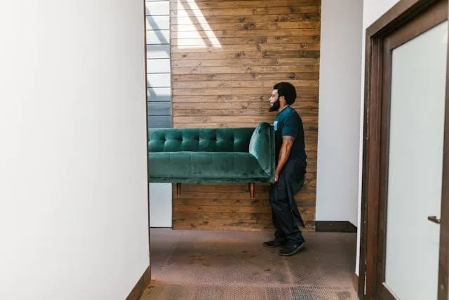 A man in jeans and a dark t-shirt carrying one half of a couch down a hallway