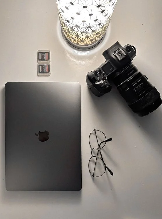Top-down flat lay of a photography workspace featuring a black DSLR-style camera with zoom lens, space gray Apple MacBook, eyeglasses, two memory cards, and a decorative lamp on a white desk.